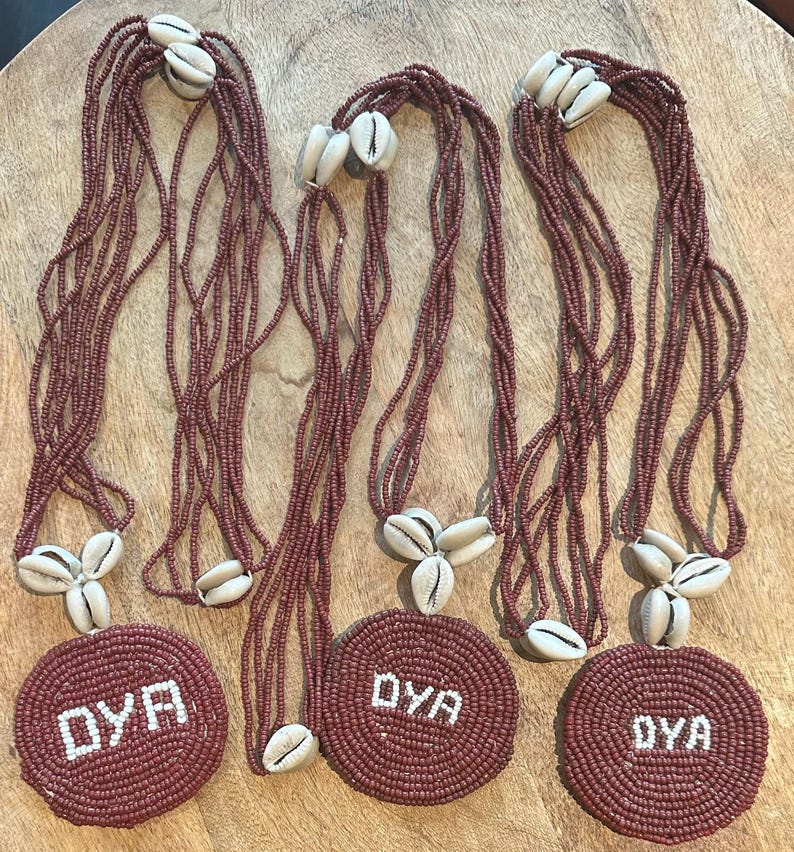 May include: Three necklaces featuring multiple strands of small, dark red beads. Each necklace has a circular pendant made of dark red beads with the white text OYA centered on it. The pendants are accented with three white cowrie shells.