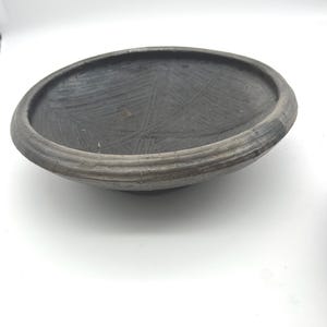 May include: A shallow, circular bowl with a dark charcoal gray finish. The interior surface features a series of intersecting, etched linear patterns. The rim is thick and rounded, showing a slightly lighter gray texture compared to the center.