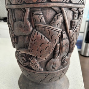 May include: A dark brown wooden vase with intricate carvings of figures and geometric patterns. The vase has a wide mouth and a narrow base.