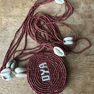 May include: A multi-strand necklace featuring small, dark reddish-brown beads and several white cowrie shells. A large, circular pendant made of the same dark reddish-brown beads displays the white text OYA in the center of the circular design.