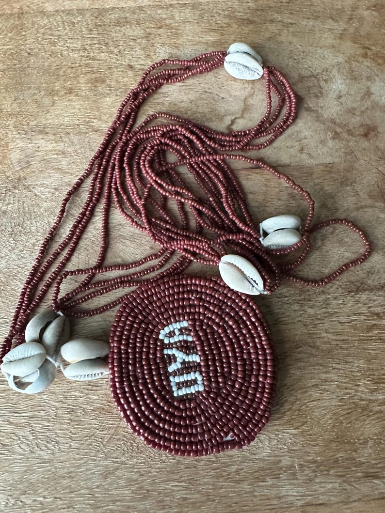 May include: A multi-strand necklace featuring small, dark reddish-brown beads and several white cowrie shells. A large, circular pendant made of the same dark reddish-brown beads displays the white text OYA in the center of the circular design.