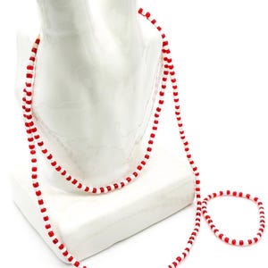 Op de afbeelding: Een lange ketting en een bijpassende armband gemaakt van kleine, afwisselend rode en witte cilindervormige kralen. De sieraden zijn geregen aan een flexibel koord en hebben een herhalend patroon van rode kralen gescheiden door witte kralen.