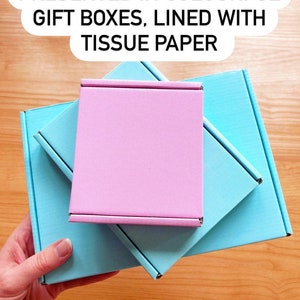 May include: Three gift boxes stacked on top of each other. The top box is pink, the middle box is light blue, and the bottom box is a darker shade of blue. The boxes are lined with tissue paper. "PRESENTED IN COLOURFUL GIFT BOXES, LINED WITH TISSUE PAPER" is written in black text on a white background.