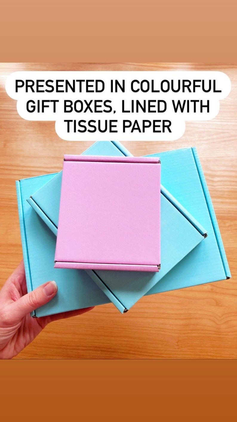 May include: Three gift boxes stacked on top of each other. The top box is pink, the middle box is light blue, and the bottom box is a darker shade of blue. The boxes are lined with tissue paper. "PRESENTED IN COLOURFUL GIFT BOXES, LINED WITH TISSUE PAPER" is written in black text on a white background.
