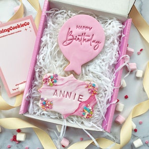 May include: A pink gift box filled with white tissue paper contains two pink cookies. One cookie is shaped like a balloon with the text "Happy Birthday" and the other cookie is a marbled pink cookie with the name "Annie" written on it.