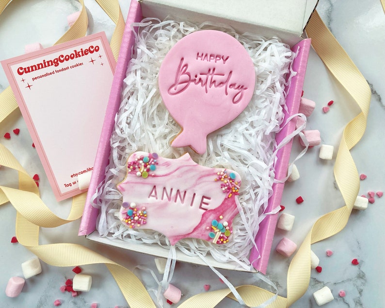 May include: A pink gift box filled with white tissue paper contains two pink cookies. One cookie is shaped like a balloon with the text "Happy Birthday" and the other cookie is a marbled pink cookie with the name "Annie" written on it.
