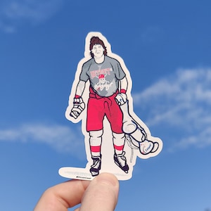 May include: A sticker depicting a cartoon hockey player in a grey Detroit jersey, red shorts, and white socks. The player is holding a hockey stick bag with the name "Cooper". The background is a blue sky.