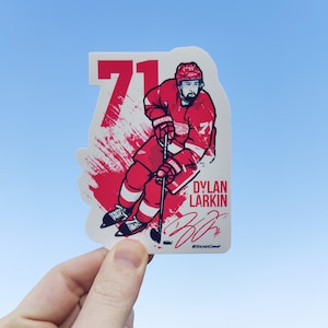 May include: A red, white, and blue hockey player sticker with the number 71 and the name Dylan Larkin. The sticker has a white background with red and white paint splatters. The player is holding a hockey stick and is in a skating position. The sticker has the text "StickerChamp" at the bottom.