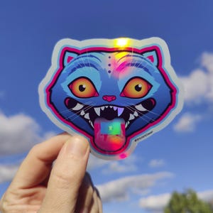 May include: A holographic sticker featuring a cartoon cat's face with a blue body, pink accents, and large yellow eyes. The cat has its tongue out, with a rainbow-colored square on the tongue. The sticker is held against a blue sky.