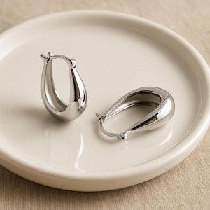 May include: Two silver-colored hoop earrings with a teardrop shape. The earrings are smooth and shiny, reflecting light. They are displayed on a white, round plate, suggesting a jewelry or accessory theme.