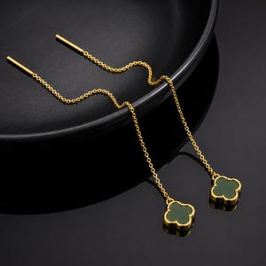 May include: A pair of gold-tone threader earrings featuring a delicate chain design that terminates in a four-leaf clover motif. Each clover charm is centered with a smooth, dark green inlay, creating a contrast against the polished gold metal.