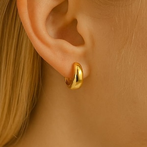 May include: A close-up of a gold-colored hoop earring. The earring is a small, rounded design, and it is worn on the ear. The earring has a shiny, polished finish.