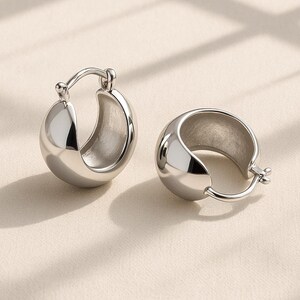 May include: Two polished silver hoop earrings. Each earring features a rounded, hollow design with a hinged clasp. The earrings are presented on a neutral surface, with gentle shadows.