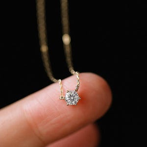May include: A delicate gold necklace featuring a single, sparkling round-cut diamond pendant. The diamond is set in a six-prong setting, and the chain is a fine, elegant design. The necklace is shown against a dark background.