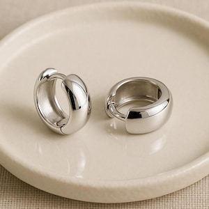 May include: A pair of shiny silver hoop earrings. The earrings are thick and rounded, with a smooth, reflective surface. They are displayed on a cream-colored, circular plate. The earrings are a simple, yet elegant design.