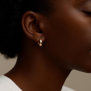 May include: A close-up of a gold hoop earring. The earring is a small, curved design, and it is worn on the ear. The earring has a smooth, polished surface and reflects light.