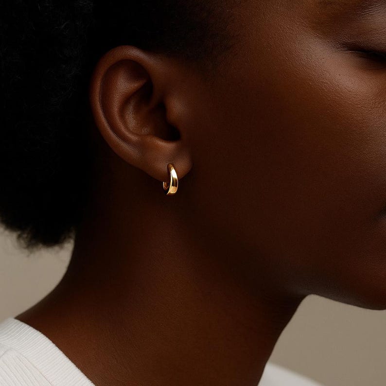 May include: A close-up of a gold hoop earring. The earring is a small, curved design, and it is worn on the ear. The earring has a smooth, polished surface and reflects light.