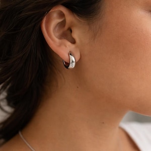 May include: A close-up of a silver hoop earring. The earring is a thick, rounded design, reflecting light. The earring is worn on the ear of a person. The background is blurred, focusing on the jewelry.