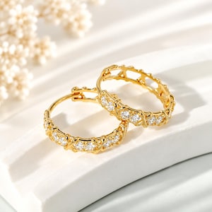 May include: A pair of gold hoop earrings featuring a repeating X-shaped metal pattern interspersed with small, clear, round-cut stones set along the front-facing surface of each circular hoop, creating a continuous sparkling design.