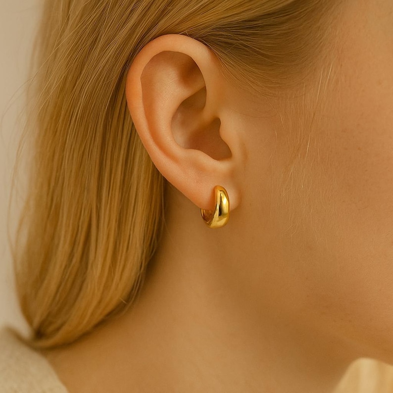 May include: A close-up of a gold-colored hoop earring. The earring is a small, rounded design, and it is worn on the ear. The earring has a shiny, polished finish.