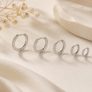 May include: A set of five silver-toned hoop earrings arranged in a row from largest to smallest. Each hoop features a row of small, clear, sparkling stones embedded along the front-facing outer edge of the circular metal band.