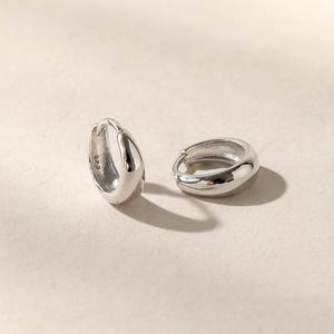 May include: A pair of small, polished silver hoop earrings with a thick, rounded profile. One earring is positioned upright, revealing a small 925 hallmark stamp on the inner surface, while the other lies flat, showcasing the smooth, reflective finish.