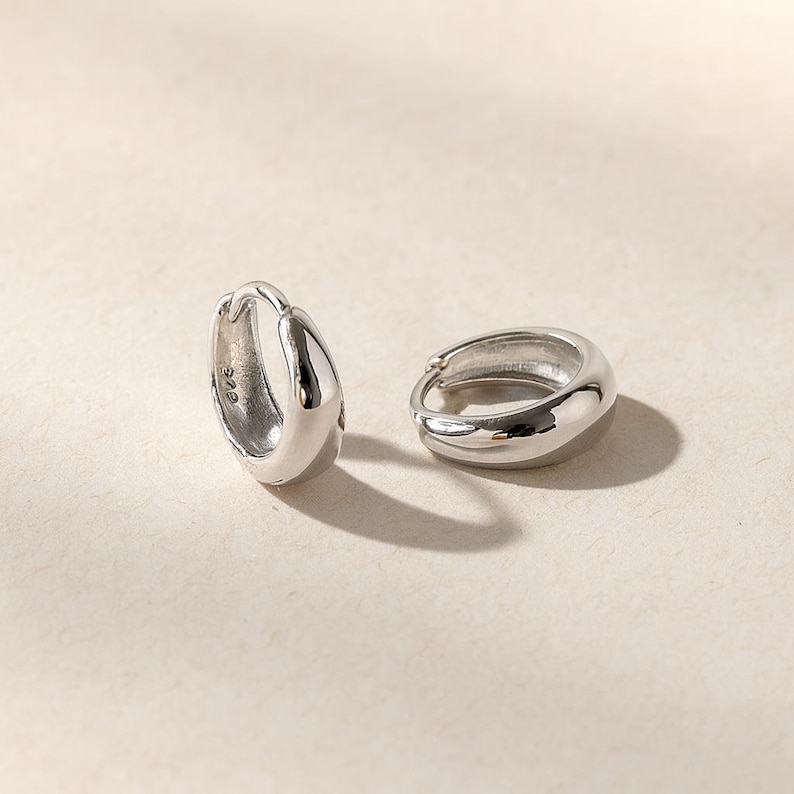 May include: A pair of small, polished silver hoop earrings with a thick, rounded profile. One earring is positioned upright, revealing a small 925 hallmark stamp on the inner surface, while the other lies flat, showcasing the smooth, reflective finish.