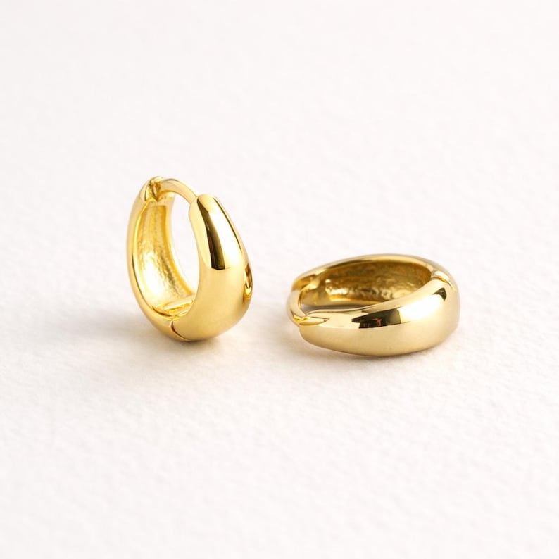 May include: Two gold-coloured hoop earrings. The earrings have a rounded, polished design. The hoops are small and appear to be made of a shiny metal. The earrings are displayed on a white background.