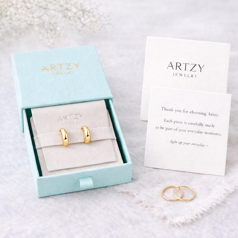 May include: A pair of gold hoop earrings presented in a light blue box with gold lettering reading "ARTZY". The box is open, showing the earrings on a beige fabric. A card with the brand name and a message is also visible.