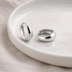 May include: Two silver hoop earrings with a smooth, rounded design. The earrings are placed on a white, slightly curved surface. One earring has the text "S925" engraved on it. The earrings are reflective and appear to be made of metal.