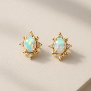 May include: A pair of gold-colored stud earrings. Each earring features an oval, iridescent opal stone surrounded by a starburst design with small, clear crystals. The earrings are set against a neutral background.