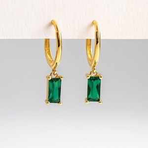 May include: A pair of gold-coloured hoop earrings, each featuring a rectangular emerald green gemstone pendant. The earrings are presented against a white backdrop, highlighting their design and colour.