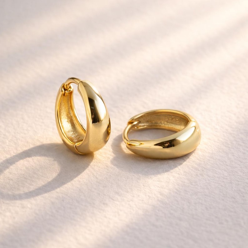 May include: A pair of gold-coloured hoop earrings. The earrings have a smooth, rounded design and a shiny finish. The earrings are displayed on a light-coloured surface, with soft shadows.