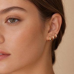 May include: Close-up of a person's ear adorned with three graduated gold hoop earrings. The earrings are smooth and shiny, reflecting light. The skin tone is fair with a soft, natural appearance. The background is a solid, neutral color.