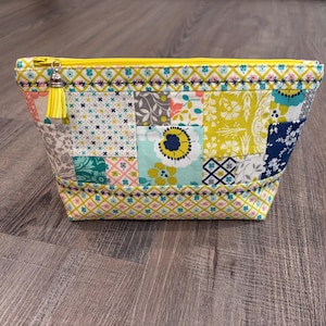 May include: A zippered pouch featuring a patchwork design of various floral and geometric patterns in shades of yellow, teal, navy blue, and grey. The bag has a yellow zipper closure with a matching yellow tassel attached to the metal pull tab.