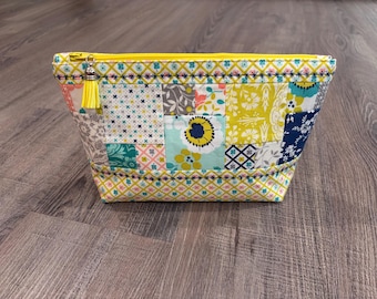 May include: A zippered pouch featuring a patchwork design of various floral and geometric patterns in shades of yellow, teal, navy blue, and grey. The bag has a yellow zipper closure with a matching yellow tassel attached to the metal pull tab.