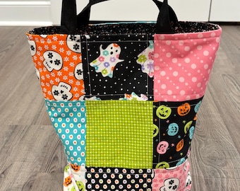 May include: A fabric tote bag featuring a patchwork design with various Halloween-themed patterns including skulls, ghosts, and pumpkins in orange, black, pink, green, and blue. The bag has two black fabric handles and a multi-colored interior lining.