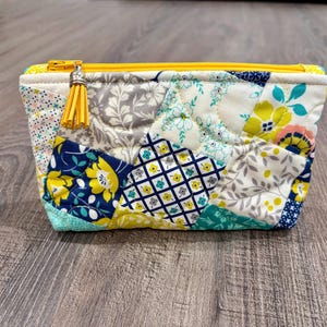 May include: A quilted zippered pouch featuring a patchwork design of various floral and geometric patterns in shades of yellow, navy blue, teal, grey, and white. The pouch has a bright yellow zipper closure and a small yellow tassel attached to the pull.