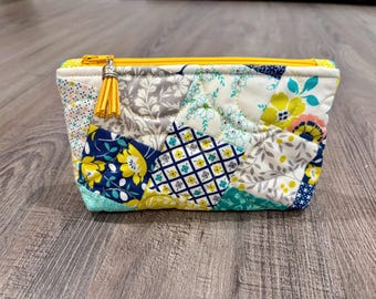 May include: A quilted zippered pouch featuring a patchwork design of various floral and geometric patterns in shades of yellow, navy blue, teal, grey, and white. The pouch has a bright yellow zipper closure and a small yellow tassel attached to the pull.