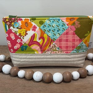 K&ouml;nnte beinhalten: Eine Rei&szlig;verschlusstasche mit einem Patchwork-Design aus bunten Blumen-, geometrischen und Karomustern in Orange, Pink, Gr&uuml;n und Blau. Der Boden besteht aus beigem Stoff mit horizontalen N&auml;hten und einem kleinen blauen Blumenanh&auml;nger.