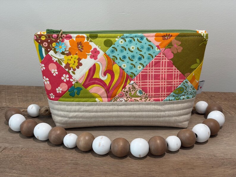 K&ouml;nnte beinhalten: Eine Rei&szlig;verschlusstasche mit einem Patchwork-Design aus bunten Blumen-, geometrischen und Karomustern in Orange, Pink, Gr&uuml;n und Blau. Der Boden besteht aus beigem Stoff mit horizontalen N&auml;hten und einem kleinen blauen Blumenanh&auml;nger.
