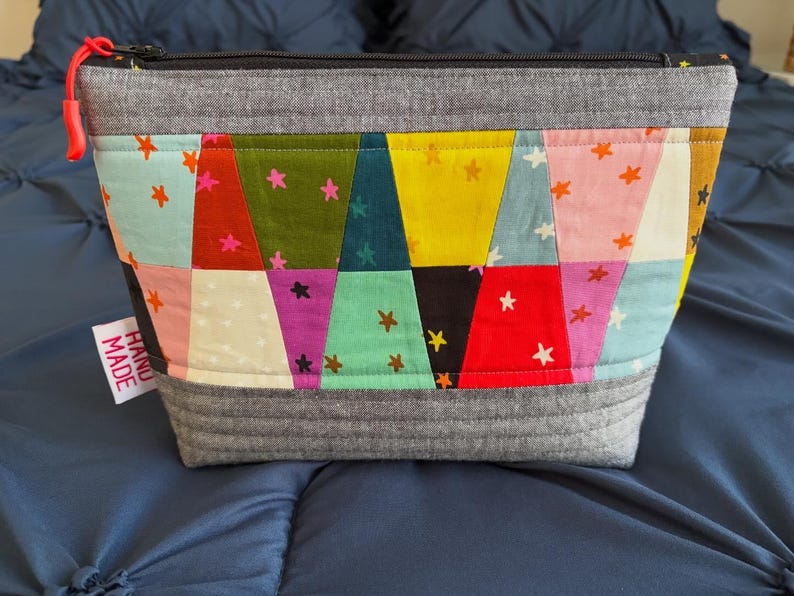 Pu&ograve; includere: Un astuccio con cerniera colorato e fatto a mano con un design patchwork. L'astuccio presenta quadrati di tessuto nei toni del rosso, rosa, verde, giallo e viola, ciascuno adornato con piccoli motivi a stella. La parte superiore e inferiore sono grigie e una cerniera rossa &egrave; visibile.