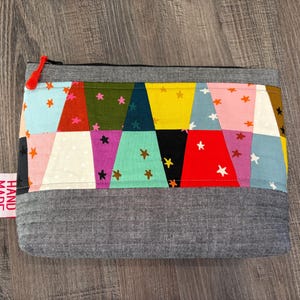 Pu&ograve; includere: Un astuccio con cerniera fatto a mano con un design patchwork. La sezione superiore presenta triangoli colorati con motivi a stella in vari colori, tra cui blu, rosa e giallo. Il fondo &egrave; in tessuto grigio. Un'etichetta rossa recita "HAND MADE".