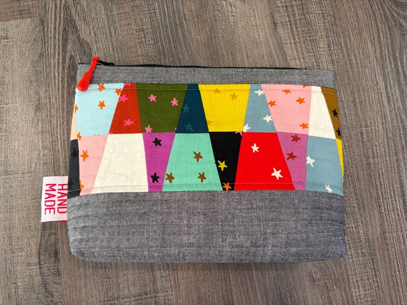 Pu&ograve; includere: Un astuccio con cerniera fatto a mano con un design patchwork. La sezione superiore presenta triangoli colorati con motivi a stella in vari colori, tra cui blu, rosa e giallo. Il fondo &egrave; in tessuto grigio. Un'etichetta rossa recita "HAND MADE".