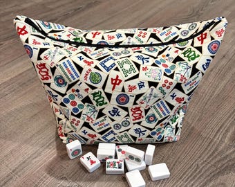 Puede incluir: Una bolsa de tela con un patrón repetitivo de fichas de Mahjong en rojo, verde, azul y negro sobre un fondo blanco, con una cremallera negra. Varias fichas de Mahjong de plástico blanco están esparcidas frente a la bolsa.