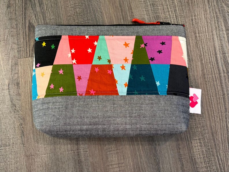 Pu&ograve; includere: Un astuccio in tessuto grigio con un colorato design patchwork. Il patchwork presenta triangoli in vari colori come rosso, rosa e verde acqua, adornati con motivi a stella bianchi e arancioni. L'astuccio ha una cerniera rossa e una piccola etichetta con un logo a cuore.