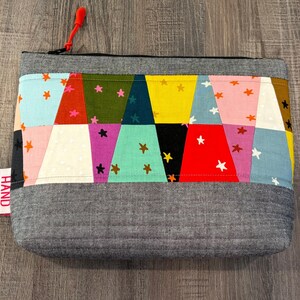Pu&ograve; includere: Una pochette rettangolare in tessuto grigio con un colorato design patchwork. Il patchwork presenta triangoli in vari colori come blu, rosa, giallo e verde, adornati da forme di stelle. Un tirazip rosso e un'etichetta "HAND MADE" sono visibili.