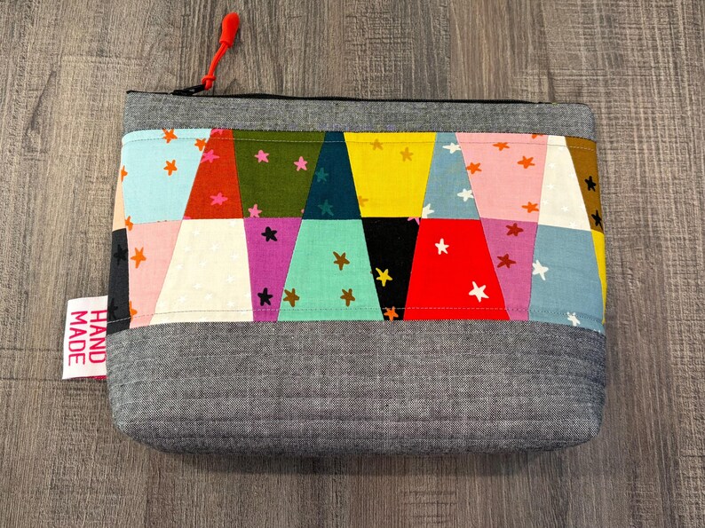 Pu&ograve; includere: Una pochette rettangolare in tessuto grigio con un colorato design patchwork. Il patchwork presenta triangoli in vari colori come blu, rosa, giallo e verde, adornati da forme di stelle. Un tirazip rosso e un'etichetta "HAND MADE" sono visibili.