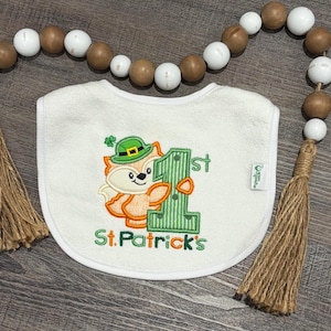 May include: White baby bib with a St. Patrick's Day theme. Features an embroidered fox wearing a green leprechaun hat next to a green number 1. The text "St. Patrick's" is embroidered in green.
