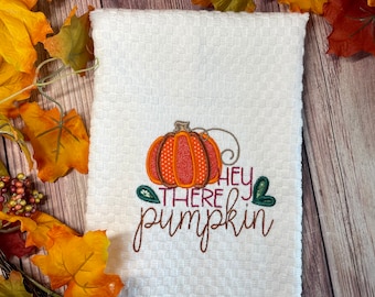 May include: White kitchen towel with a textured pattern, featuring an embroidered orange pumpkin with a dotted design and the phrase "Hey There Pumpkin" in brown script. Autumn leaves surround the towel.